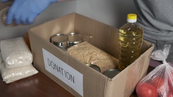 εθελοντής φορώντας προστατευτικά γάντια βάζοντας τρόφιμα παντοπωλείου στο κουτί δωρεάς χαρτοκιβωτίων - Πλάνα, βίντεο
