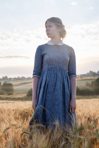 young girl in blue dress with collected hair posing with her eyes closed in field at sunrise. Foggy dawn in background - Photo, Image
