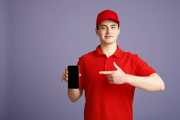 Cute courier in red uniform points finger at smartphone with blank screen, isolated gray background - Foto, Imagem