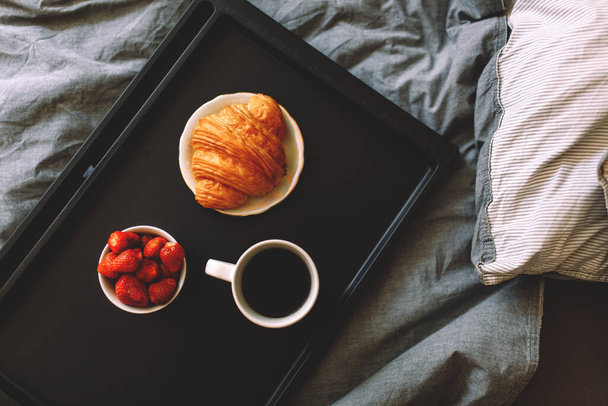 Breakfast on a tray in bed at home white dark linen. croissant tea strawberries - Foto, afbeelding