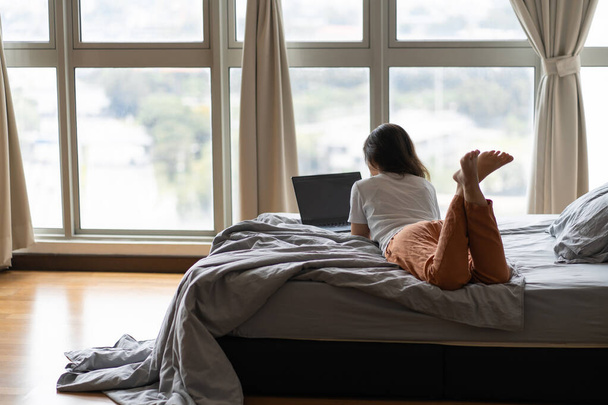 A beautiful young brunette girl is working on the laptop while lying on a bed by a panoramic window with beautiful view from a high floor. Stylish modern interior. A cozy workplace. Shopping Internet. - Photo, Image