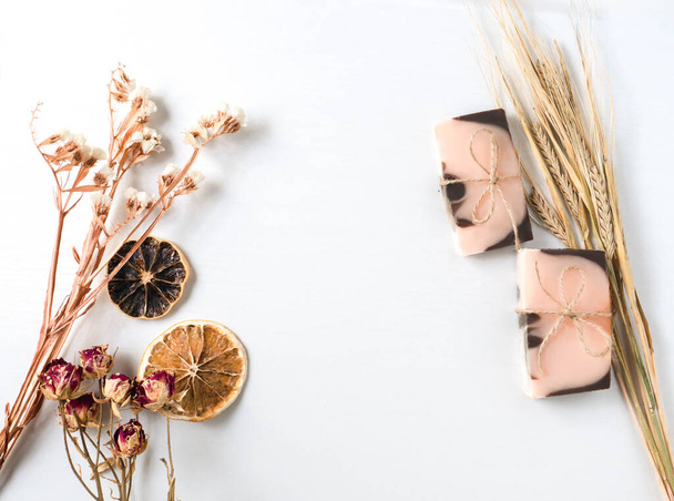 Flat lay composition with two handmade soaps of brown-pink color with an orange tint, dried slices of oranges, roses flowers and spikes of wheat on a white background with wooden texture. Horizontal - Fotó, kép