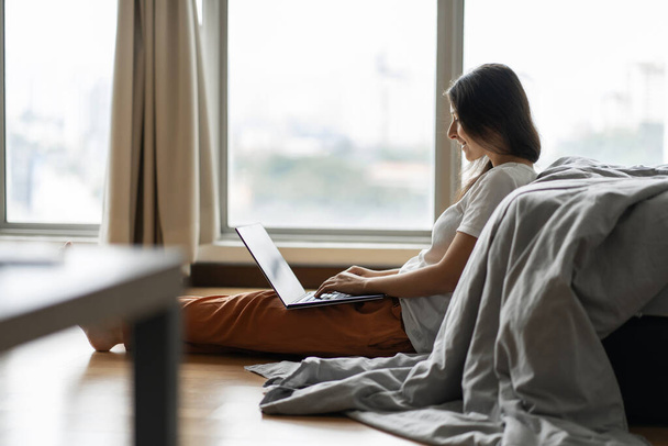 Beautiful young brunette girl working on a laptop, sitting on the floor near the bed by the panoramic window with a beautiful view from the high floor. Stylish modern interior.Shopping Internet - Φωτογραφία, εικόνα