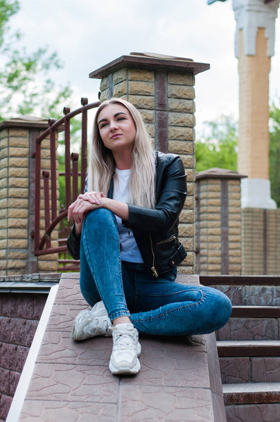 Cute positive blonde girl in a black leather jacket and blue jeans in the spring on the porch, near a beautiful house - Photo, Image