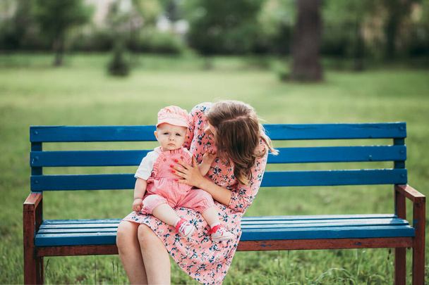ピンクの服を着た公園のベンチに座っている小さな子供を持つ若い美しい母親. - 写真・画像
