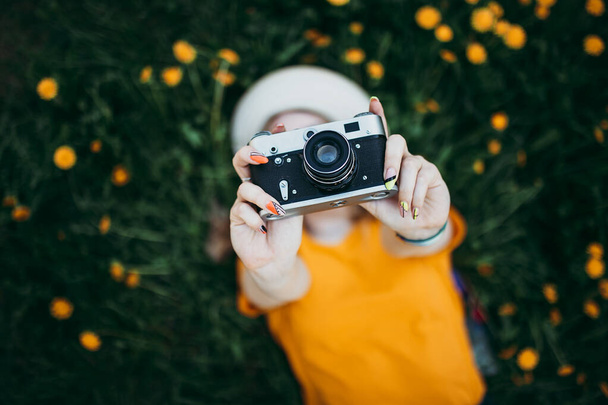Junges schönes Mädchen in orangefarbenem T-Shirt mit Retro-Kamera in der Hand auf dem Rasen liegend, wo Löwenzahn wächst, blühender Löwenzahn. - Foto, Bild