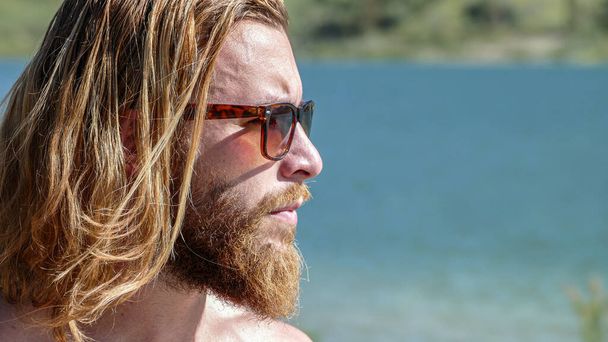Blond młody człowiek w okularach przeciwsłonecznych patrząc na horyzont jeziora - Zdjęcie, obraz