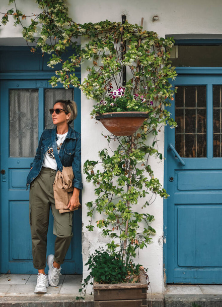 Pretty fashionable woman standing in doorway of old house. Cool dressed lady in front of turquoise wooden door with flowers around. - Photo, Image