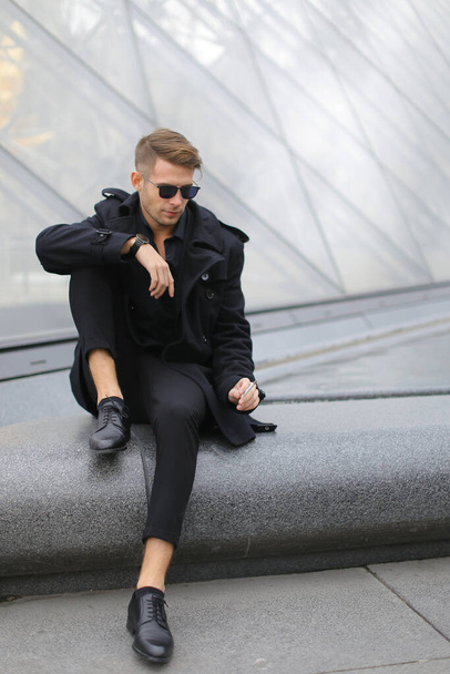 Jonge Europeaan in zwart jasje zittend op Louvre Pyramide en roken. - Foto, afbeelding