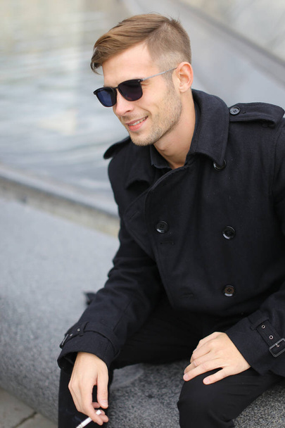 若いです笑顔ブロンド男身に着けている黒いジャケットに座っているルーブルピラミッドと喫煙. - 写真・画像