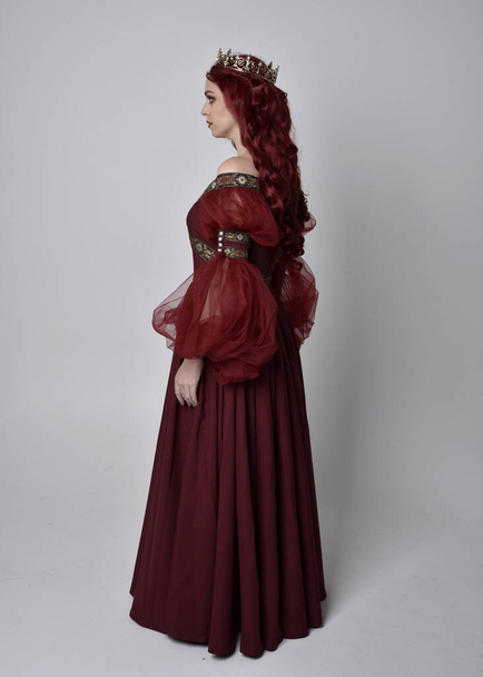 Портрет красивої жінки з рудим волоссям у бургундській фентезійній сукні та золотій короні. Поза довжиною, ізольована на фоні студії. - Фото, зображення