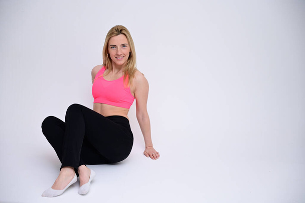 Portrait of a pretty fitness blonde girl in a sports uniform on a white background sitting on the floor. A slender Caucasian model poses with a smile. - Foto, Imagen