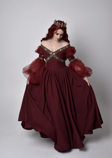 Portret van een mooie vrouw met rood haar met een vloeiende Bourgondische fantasiejurk en gouden kroon. staande houding op volledige lengte, geïsoleerd tegen een studioachtergrond - Foto, afbeelding