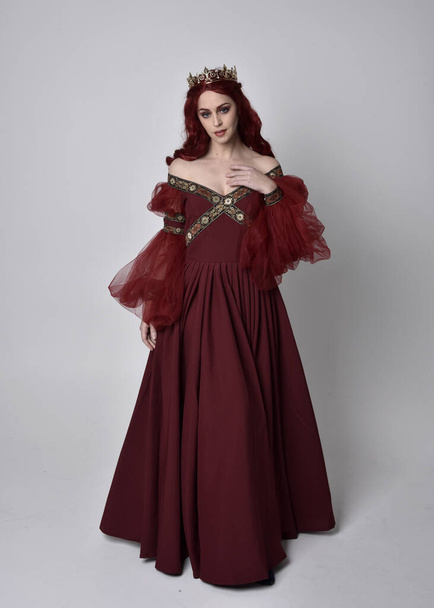 Retrato de uma mulher bonita com cabelo vermelho vestindo um vestido de fantasia Borgonha fluindo e coroa de ouro. postura ereta de comprimento total, isolada contra um fundo de estúdio
 - Foto, Imagem