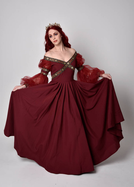 Portret van een mooie vrouw met rood haar met een vloeiende Bourgondische fantasiejurk en gouden kroon. staande houding op volledige lengte, geïsoleerd tegen een studioachtergrond - Foto, afbeelding