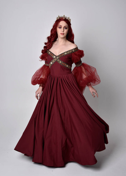 Portrait of a beautiful woman with red hair wearing  a  flowing Burgundy fantasy gown and golden crown.  full length standing pose, isolated against a studio background - Photo, Image