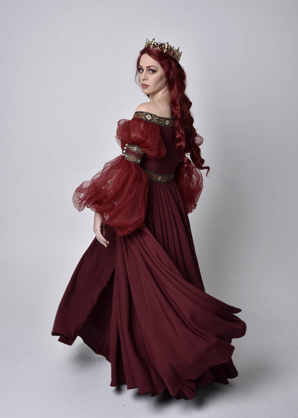 Portret pięknej kobiety z rudymi włosami ubranej w płynącą burgundzką suknię fantazyjną i złotą koronę. pełnowymiarowa pozycja stojąca, izolowana na tle studia - Zdjęcie, obraz