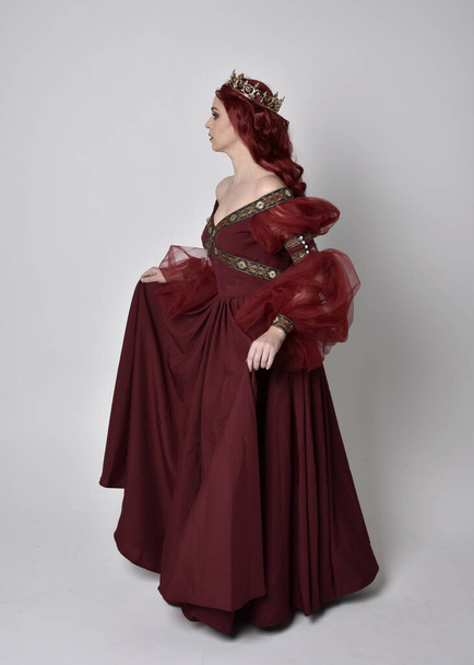Портрет красивой женщины с рыжими волосами в бургундском фэнтези-платье и золотой короне. полностью стоящая поза, изолированная от студийного фона
 - Фото, изображение