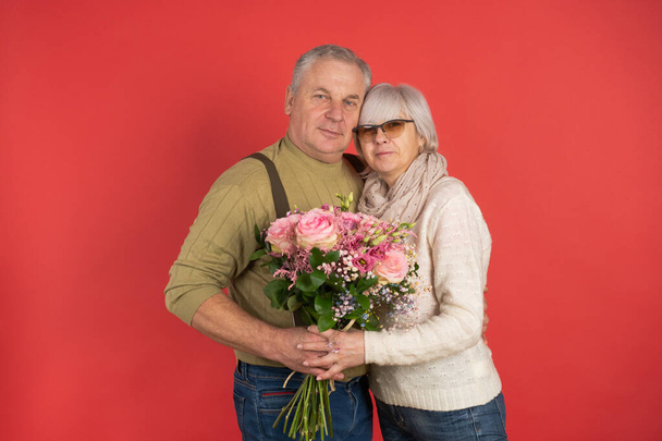 An elderly man gives a beautiful bouquet of flowers to an elderly woman, wife, colleague, friend, red background, the concept of happy old age - Photo, Image