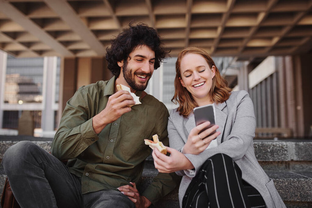 Couple watching video on smartphone while eating sandwich during break from office - Foto, Imagem