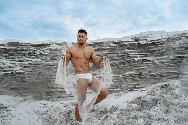 sexy macho hombre en pantalones cortos blancos se sienta cerca en la arena contra el fondo azul cielo sostiene la arena en sus manos despertar a través de los dedos, fitness culturista iluminado por el sol
 - Foto, Imagen