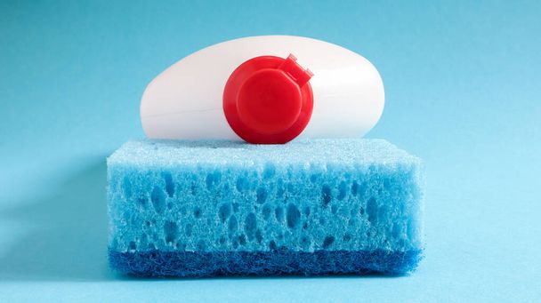 Een blauwe spons met een witte fles wasmiddel op een blauwe achtergrond. Gemak van sponzen bestaat uit goede detergent retentie die het mogelijk maakt om het economisch te gebruiken. - Foto, afbeelding