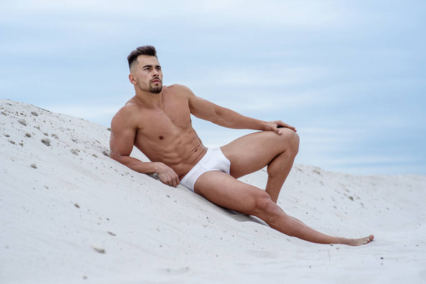 sexy man, white macho macho man lying on white sand against sky background, white empty beach without people, athlete sunbathes on beach - Photo, Image