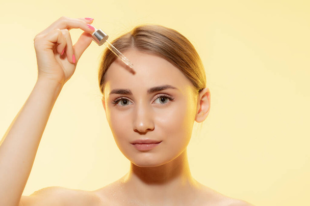 Protection. Close up of beautiful female face with pouring essential oil, serum over yellow background. Cosmetics and makeup, natural and eco treatment, skin care. - Foto, Imagem
