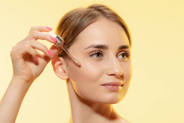 Protection. Close up of beautiful female face with pouring essential oil, serum over yellow background. Cosmetics and makeup, natural and eco treatment, skin care. - Photo, Image
