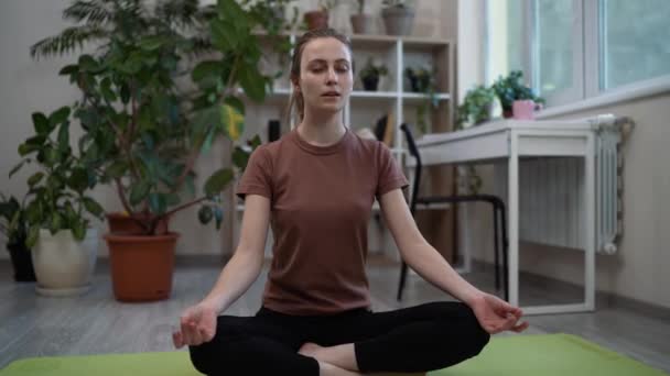 Quédate en casa, mantente en forma. Hermosa mujer joven tranquila meditando, haciendo ejercicios de yoga mientras está sentada en la pose de loto en el suelo en casa
 - Metraje, vídeo