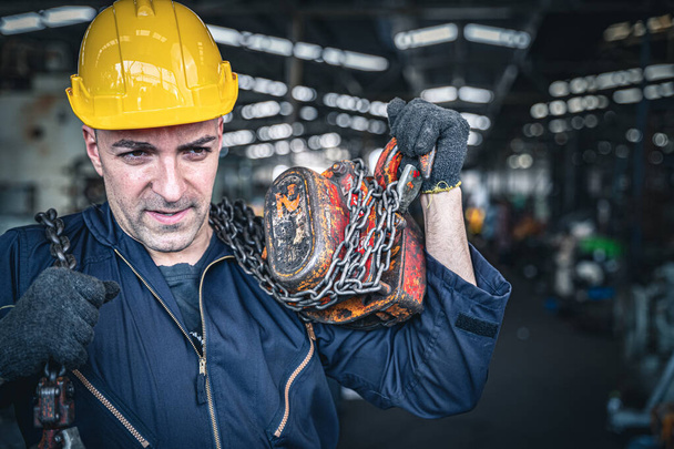 Trabajador industrial masculino en mono azul y polipasto de cadena de carga de casco duro en su espalda
. - Foto, Imagen