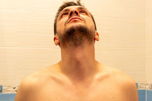 young attractive shirtless man sitting in the bathroom on the background of a light tile and looking up. - 写真・画像