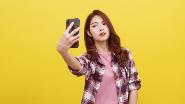 Hymyilevä suloinen aasialainen nainen tekee selfie-valokuvan älypuhelimella, jolla on positiivinen ilme rennoissa vaatteissa ja katsomalla kameraa keltaisella taustalla. Onnellinen ihana iloinen nainen iloitsee menestyksestä. - Materiaali, video