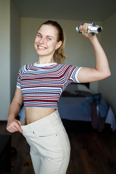 Smiling slim woman in big pants with dumbbell in hand in apartment. - Foto, Bild