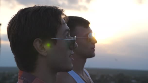 Profil młodej pary męskiej w okularach przeciwsłonecznych stojącej na krawędzi dachu i mówiącej. Przystojni geje relaksujący się na dachu wieżowca i cieszący się pięknym krajobrazem miejskim. Zamknij Powolny ruch - Materiał filmowy, wideo