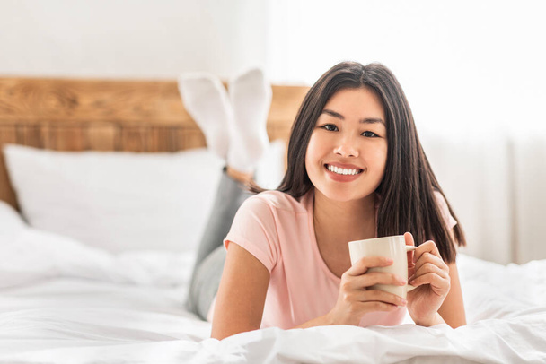 Asyalı kız sıcak yatak odasında uzanıp kahve içiyor. - Fotoğraf, Görsel