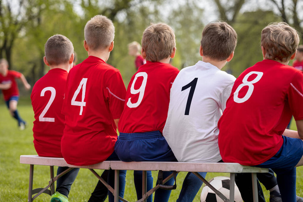 Equipa Sports Background. Rapazes numa equipa desportiva sentados juntos num banco. Crianças vestindo camisas de futebol vermelho com números nas costas. Nível Escolar Futebol Equipe Jogando Match
 - Foto, Imagem
