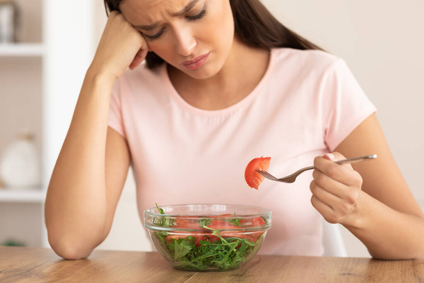 Θλιβερή γυναίκα τρώει σαλάτα αδυνατίσματος κάθεται στο τραπέζι στο σπίτι - Φωτογραφία, εικόνα