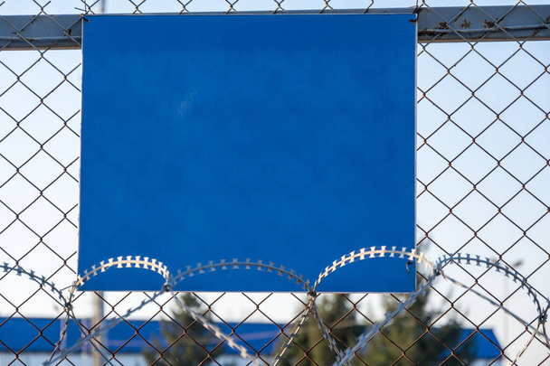 Close-up view of a caution banner, mock-up of a blue plate on an iron mesh fence with barbed wire. Closed area designation blank. Quarantine and lockdown concept - Photo, Image