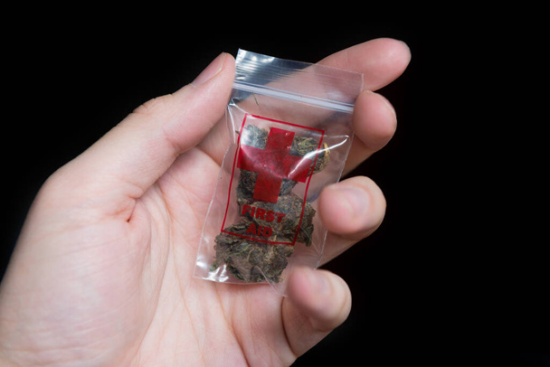 mão masculina segurando pequeno saco poli com impressão de primeiros socorros e maconha medicinal nele, isolado em fundo preto
 - Foto, Imagem