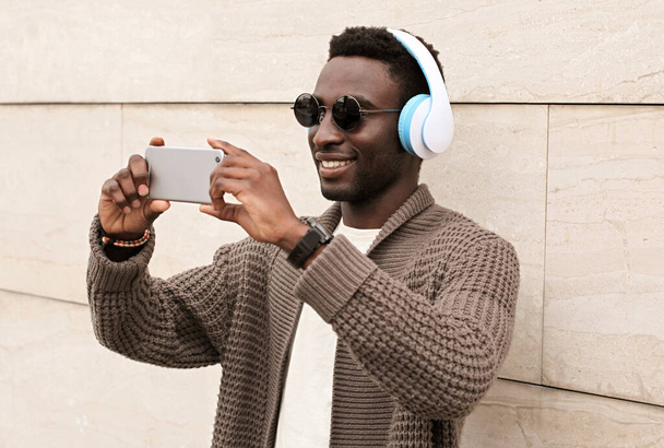 Portrait stylish smiling african man taking selfie picture by smartphone in wireless headphones listening to music wearing brown knitted cardigan, sunglasses on city street over wall background - Foto, imagen