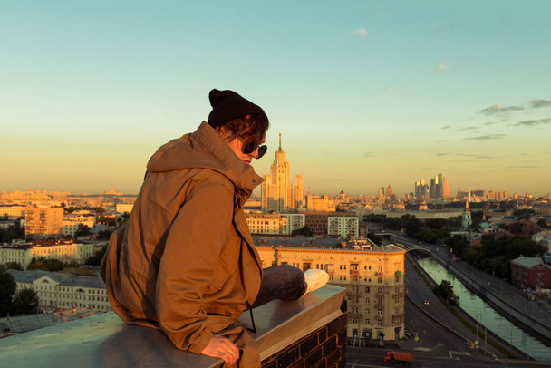 MOSCOU, RUSSIE - 13 JUIN 2016 : Jeune homme courageux assis sur le bord d'un toit élevé. Moscou ville en été à l'aube sur fond
 - Photo, image