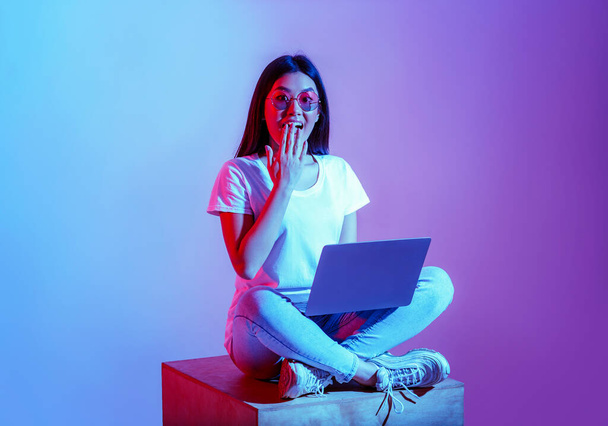 Σύγχρονη blogger με φορητό υπολογιστή στο χώρο εργασίας. Έκπληκτη κοπέλα με γυαλιά κλείνει το στόμα με το χέρι - Φωτογραφία, εικόνα