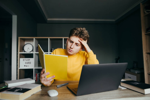 Красивий молодий чоловік в жовтому светрі читає блокнот, сидячи за столом вдома в спальні. Підліток вивчає дистанційне навчання вдома, використовуючи ноутбук
. - Фото, зображення