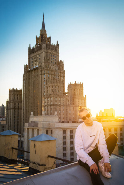 MOSCOW, RUSSIA - JUNE 30, 2016: Young and brave male sitting on the edge of high roof Ministry of Foreign Affairs building on background  - Foto, imagen