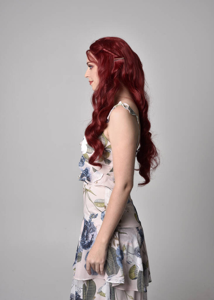 Retrato de uma mulher bonita com cabelo vermelho vestindo um vestido floral fluindo. 3 / 4 pose, isolado contra um fundo de estúdio
 - Foto, Imagem