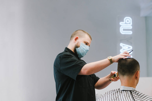Professional barber in a medical gauze mask on his face making a hairstyle for a client during quarantine. Work of the barbershop in the coronavirus, the use of personal protective equipment. - Photo, Image
