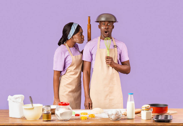 Boos zwart meisje met rollende pin en haar bang vriendje proberen om samen te bakken op kleur achtergrond - Foto, afbeelding