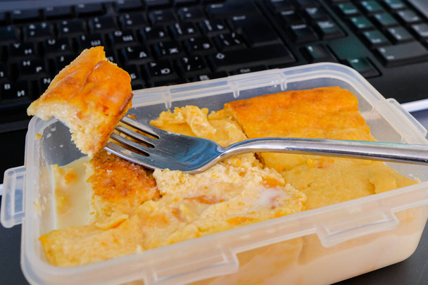 Appetizing cottage cheese casserole with pumpkin in a plastic container and a metal fork on a black computer keyboard background. Lunch at the office. Food brought with you from home. - Photo, image