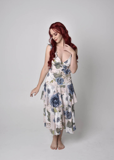 Retrato de uma mulher bonita com cabelo vermelho vestindo um vestido floral fluindo. postura ereta de comprimento total, isolada contra um fundo de estúdio
 - Foto, Imagem
